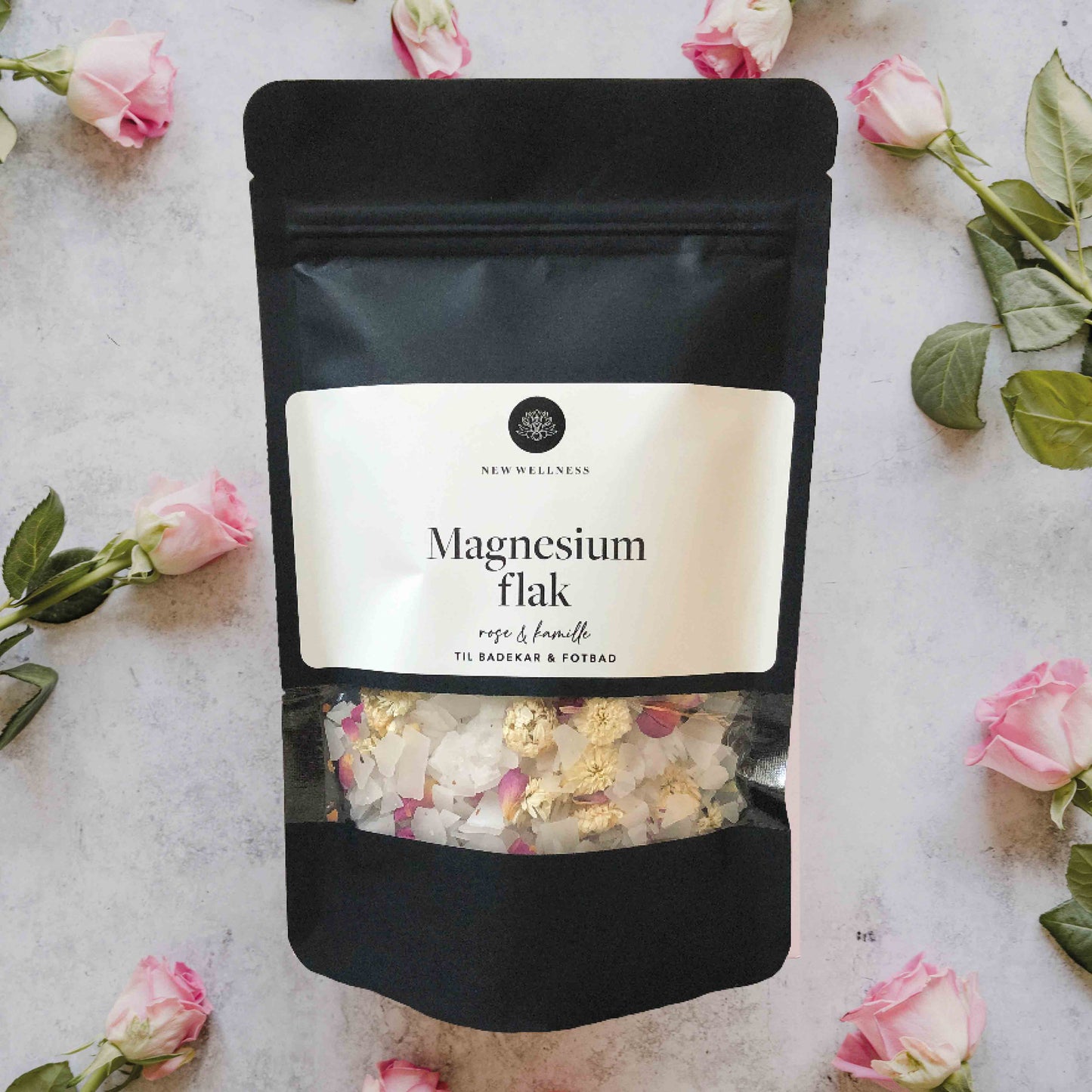 Sort pose med hvit etikett og gjennomsiktig vindu, med magnesiumflak med rose og kamille. Det er roser i bakgrunnen.