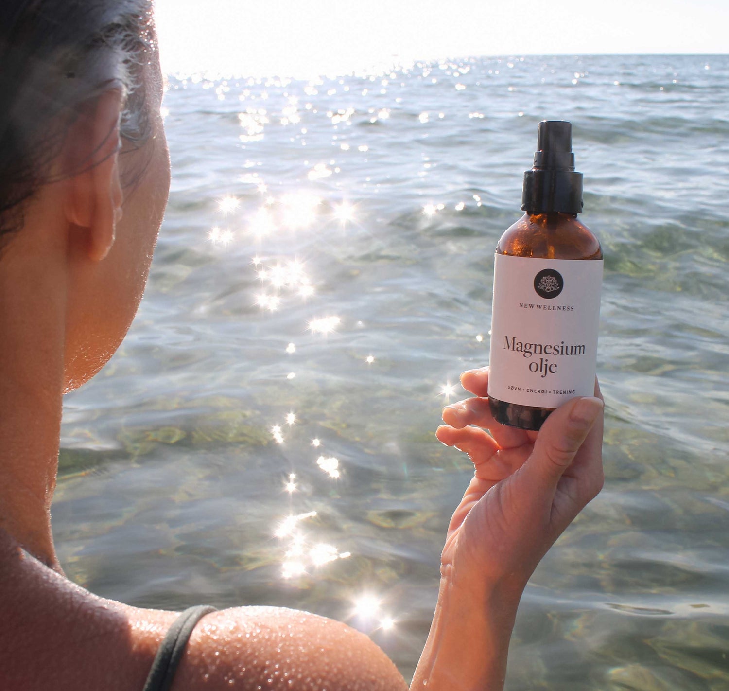 Kvinne står i et glitrende, krystallklart vann og holder en flaske magnesiumolje